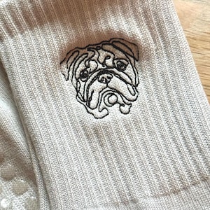 May include: A pair of light beige ribbed socks featuring a black embroidered outline of a bulldog face on the side. The fabric has a textured knit pattern, and the underside of the socks includes small, clear, circular silicone grip dots.