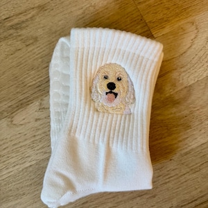 May include: A pair of white ribbed crew socks featuring an embroidered golden-colored dog face with a black nose and pink tongue. The soles of the socks have small, white, circular silicone grip dots for traction on smooth surfaces.