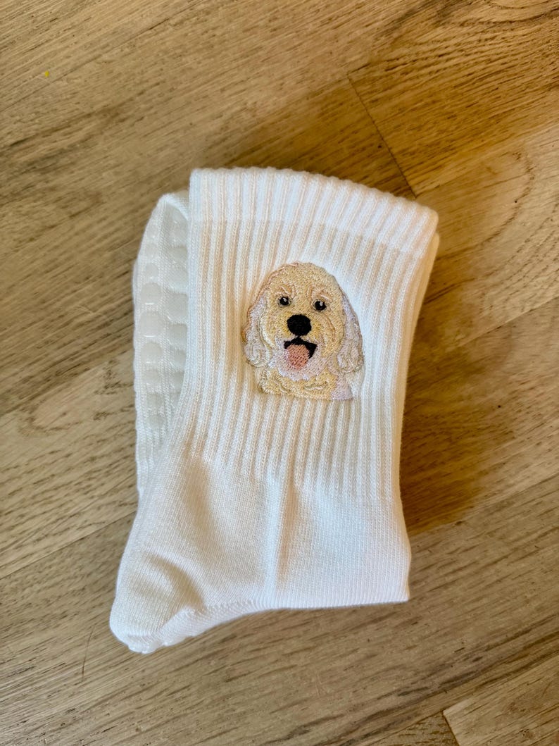 May include: A pair of white ribbed crew socks featuring an embroidered golden-colored dog face with a black nose and pink tongue. The soles of the socks have small, white, circular silicone grip dots for traction on smooth surfaces.