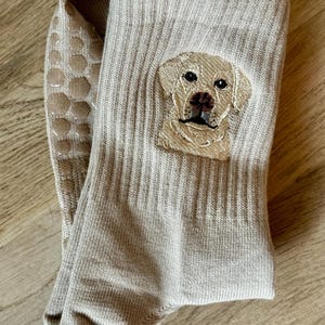 May include: A pair of beige ribbed crew socks featuring an embroidered portrait of a yellow Labrador retriever on the outer side. The soles of the socks are equipped with a series of circular, light-colored silicone grip dots for traction.