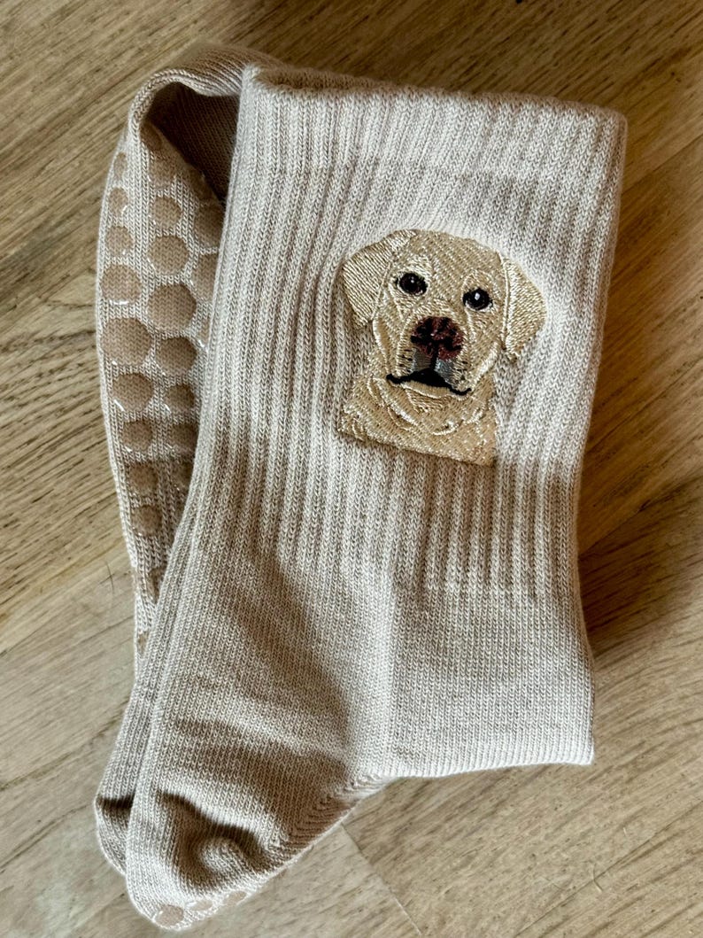 May include: A pair of beige ribbed crew socks featuring an embroidered portrait of a yellow Labrador retriever on the outer side. The soles of the socks are equipped with a series of circular, light-colored silicone grip dots for traction.