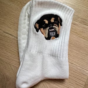 May include: A white crew sock featuring an embroidered patch of a black and tan dog face with a short snout. The sock has a ribbed cuff and a textured, white silicone grip pattern on the sole for traction, providing a non-slip surface.