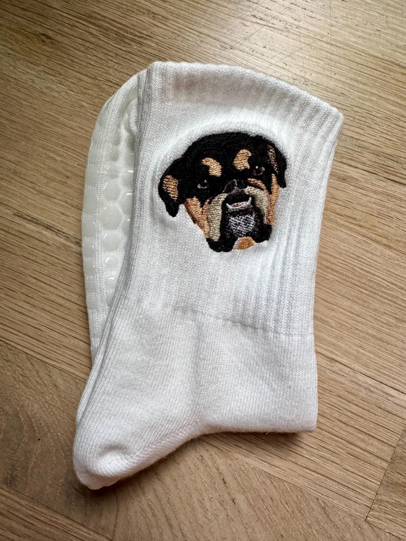 May include: A white crew sock featuring an embroidered patch of a black and tan dog face with a short snout. The sock has a ribbed cuff and a textured, white silicone grip pattern on the sole for traction, providing a non-slip surface.