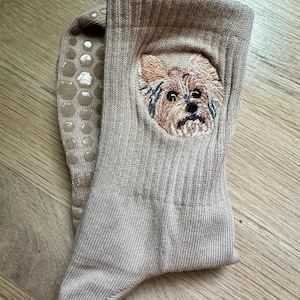May include: A pair of beige ribbed crew socks featuring an embroidered portrait of a Yorkshire Terrier on the front. The underside of the socks includes a pattern of circular, light-colored silicone grip dots to provide traction on smooth surfaces.