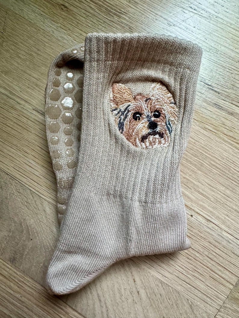 May include: A pair of beige ribbed crew socks featuring an embroidered portrait of a Yorkshire Terrier on the front. The underside of the socks includes a pattern of circular, light-colored silicone grip dots to provide traction on smooth surfaces.