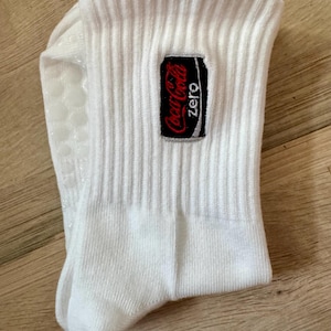 May include: A white ribbed crew sock featuring a rectangular embroidered patch with the Coca-Cola Zero logo in red, white, and black. The sole of the sock includes small, white, circular silicone grip dots for traction and stability.