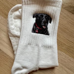 May include: A pair of white ribbed crew socks featuring an embroidered black dog with a red collar and a small bone-shaped tag. The soles of the socks have white silicone grip dots for traction, providing a textured surface on the bottom area.