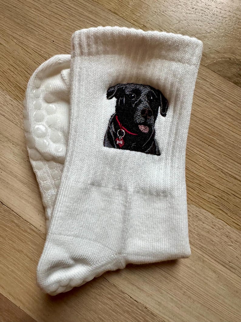 May include: A pair of white ribbed crew socks featuring an embroidered black dog with a red collar and a small bone-shaped tag. The soles of the socks have white silicone grip dots for traction, providing a textured surface on the bottom area.