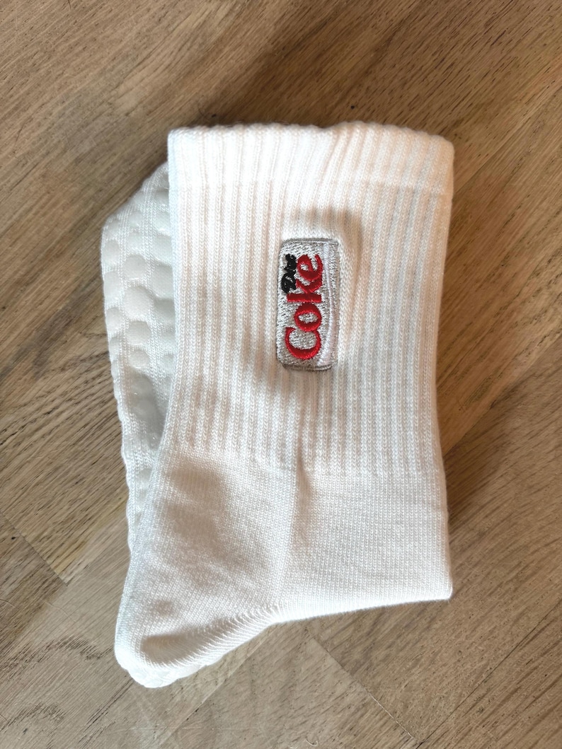 May include: White ribbed crew socks with an embroidered Coke logo in red and black. The socks are folded and displayed on a light brown wooden surface. The socks are made of a soft, comfortable material.