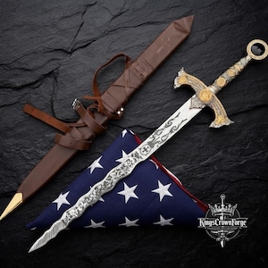 Puede incluir: Una espada de hoja ondulada plateada con una empuñadura ornamentada en oro y plata, acompañada de una vaina de cuero marrón con correas y una bandera estadounidense plegada. La hoja presenta grabados intrincados y un símbolo de cruz.