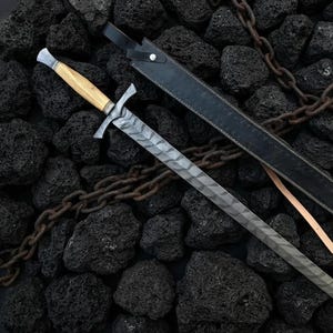 May include: A sword with a patterned steel blade, a light-coloured wooden handle, and a dark metal crossguard. The sword is accompanied by a black leather scabbard with a silver-coloured stud. The KingsCrownForge logo is visible in the corner.