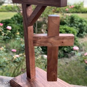 May include: A dark brown wooden sculpture featuring a vertical post with a horizontal arm and a diagonal support brace, connected to a separate smaller wooden cross that appears to be suspended in mid-air above a rectangular wooden base.