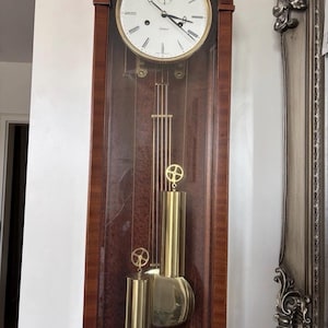 May include: A tall, dark wood grandfather clock with an arched top and a glass-fronted case. The white clock face features black Roman numerals and hands. Inside, a brass pendulum and two cylindrical brass weights hang from thin metal rods.