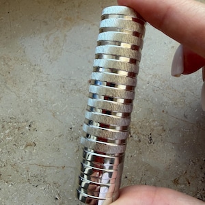 May include: A cylindrical metal object with a repeating pattern of alternating brushed silver bands and polished silver rings. The surface features a textured finish on the wider sections and smooth, reflective edges on the thinner, recessed rings.