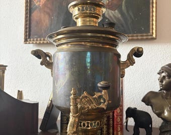 Könnte beinhalten: Ein metallischer Samowar mit einem dunklen, angelaufenen zylindrischen Körper, verzierten Messinggriffen, einem dekorativen Zapfhahn mit schwarzem Knopf und einem Sockel. Der obere Teil umfasst einen runden Deckel mit einem Messing-Kaminaufsatz.