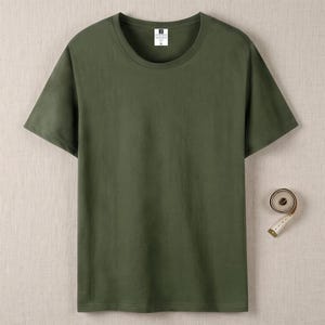 Könnte beinhalten: Ein einfarbiges, olivgrünes Kurzarm-T-Shirt mit Rundhalsausschnitt und einem weißen Innenetikett in Größe XL. Ein aufgerolltes weißes Maßband liegt rechts neben dem T-Shirt und zeigt eine Messung von 2,5 cm an.