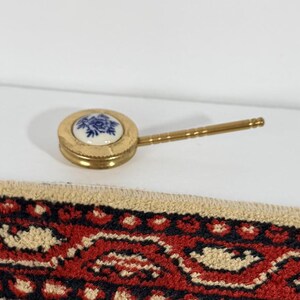 May include: A small gold-toned magnifying glass with a circular white porcelain inlay featuring a blue floral design, positioned above a rectangular woven textile mat with a red, black, and beige geometric pattern and fringed edges.