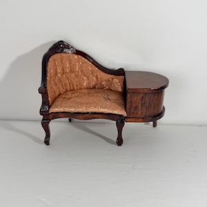 Peut inclure: Une miniature de chaise longue en bois avec une finition brun foncé et un dossier incurvé. L'assise et le dossier sont recouverts d'un tissu texturé de couleur cuivre clair. Le meuble présente une extension latérale plate et arrondie.