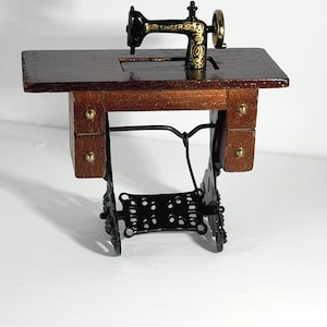 Könnte beinhalten: Eine Miniatur-Nähmaschine mit schwarzem Metallgehäuse, goldenem Singer-Schriftzug und dekorativen Verzierungen, montiert auf einem dunkelbraunen Holztisch mit zwei kleinen Schubladen und einem schwarzen Metallgestell mit kunstvollen Ausschnitten.