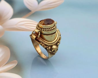 May include: A gold-toned metal ring featuring a hinged locket compartment topped with an oval amber-colored gemstone. The band is decorated with small skull motifs and beaded detailing, with a secure latch mechanism on the side of the locket.