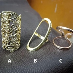 May include: Three metal rings labeled A, B, and C. Ring A is a tall, gold-tone band with an intricate, openwork filigree scroll pattern. Ring B is a smooth, elongated gold-tone oval band. Ring C is a rose gold-tone band with a split, open design.