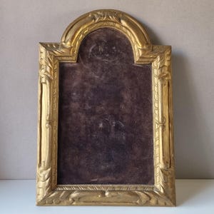 Può includere: Una cornice rettangolare con arco superiore arrotondato, caratterizzata da una finitura oro metallizzato con dettagli decorativi intagliati lungo i bordi. Il centro della cornice è riempito con un inserto in velluto marrone scuro.