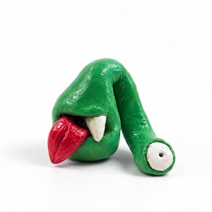 May include: A green sculpted creature figure with a glossy finish, featuring a large red tongue protruding from its mouth, a single sharp white tooth, and a long tail ending in a round white eye with a small black pupil centered on the surface.