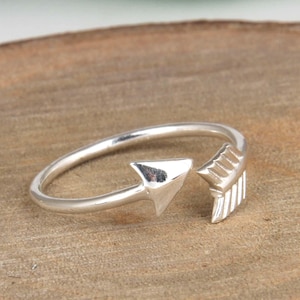 May include: A silver-toned open ring featuring an arrow design. One end of the band is shaped like a pointed arrow head, while the opposite end is shaped like the fletching or tail feathers of an arrow. The band is smooth and polished.