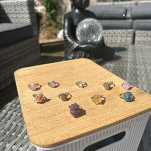 Könnte beinhalten: Eine Sammlung von zehn Ringen mit rohen, ungeschliffenen Edelsteinen in verschiedenen Farben wie Rosa, Blau, Gelb, Violett und Transparent, die jeweils auf einem dünnen silberfarbenen Metallring sitzen und auf einem hellen Holzdeckel liegen.