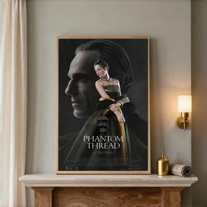 May include: A rectangular movie poster for Phantom Thread in a wooden frame. The poster features a large, shadowed profile of a man behind a woman in a dark green strapless gown. The text reads Phantom Thread, Daniel Day-Lewis, Lesley Manville, Vicky Krieps.