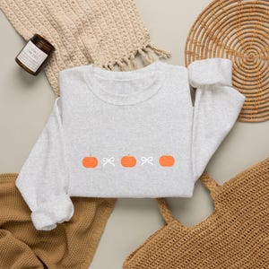 Pode incluir: Uma camisola de gola redonda cinzenta clara com um design bordado no peito. O padrão consiste em três abóboras cor de laranja alternadas com dois pequenos laços brancos, dispostos numa linha horizontal no centro da peça de vestuário.