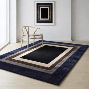 May include: A rectangular area rug featuring a nested border design with a solid black center, surrounded by concentric bands of cream, brown, gray, and deep navy blue. A light-colored wooden chair with a curved backrest sits on the rug.