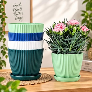 May include: A stack of four ribbed plant pots in light green, blue, white, and dark teal, accompanied by a separate light green pot containing a flowering plant with pink carnations. Each pot includes a matching saucer base for drainage.