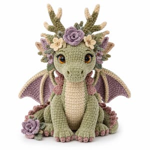 May include: A crocheted dragon plush toy in shades of sage green, cream, and dusty purple. The creature features large brown eyes, antlers, and wings, adorned with crocheted flowers on its head and one flower resting at its base on the floor.