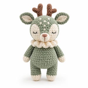 May include: A crocheted deer plush toy featuring a sage green body with cream-colored accents on the face, ears, hands, and feet. The deer has brown antlers, closed eyes with black stitched eyelashes, and a ruffled cream collar around its neck.