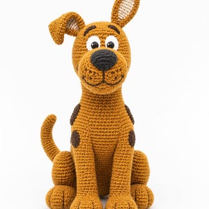 May include: A brown crocheted dog figurine with dark brown spots, a black nose, and large white eyes with black pupils. The dog has one ear standing upright and one ear folded over, a long tail, and a stitched mouth on a light brown muzzle.