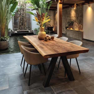 Könnte beinhalten: Ein rechteckiger Esstisch mit einer natürlichen Baumkanten-Holzplatte und schwarzen V-förmigen Metallbeinen. Vier gepolsterte beige Stühle mit schwarzen Metallbeinen stehen um den Tisch. Eine strukturierte beige Vase mit gelben und roten Blumen steht darauf.