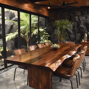 May include: A large rectangular dining table with a natural live edge wood grain surface and a thick pedestal base, accompanied by several chairs featuring dark brown wood-slat backs and black metal frames arranged in a spacious indoor setting.