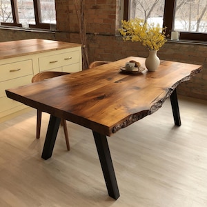 Puede incluir: Una mesa de comedor rectangular con un tablero de madera de borde natural con un acabado marrón cálido y veta visible. La mesa está sostenida por cuatro patas metálicas negras en ángulo. Un jarrón blanco con flores amarillas sobre la mesa.