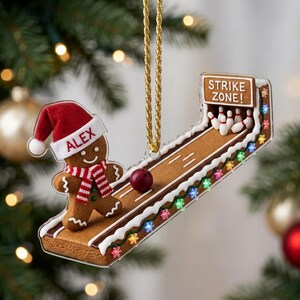 May include: A gingerbread person ornament wearing a red and white Santa hat with the name ALEX printed on it and a red and white striped scarf. The ornament features a bowling lane with a red ball, white pins, and a sign reading STRIKE ZONE!