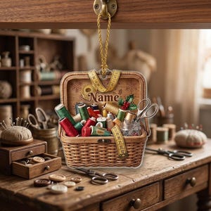 May include: A hanging ornament shaped like a woven sewing basket filled with colorful thread spools, a measuring tape, and scissors. The interior lid features the word Name, snowflake patterns, and a holly sprig. It hangs from a gold cord.