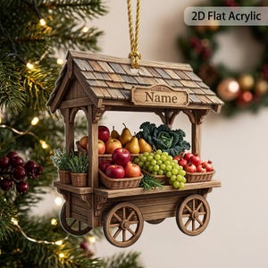 May include: A 2D flat acrylic hanging ornament shaped like a wooden market cart with a shingled roof. The cart features a sign reading Name and is filled with miniature baskets of red apples, green grapes, pears, and a head of green cabbage.