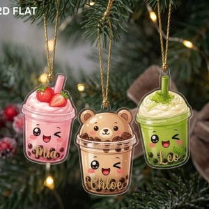 May include: Three flat hanging ornaments shaped like boba tea cups. The left is pink with strawberries and the name Mia. The center is a brown bear cup with the name Chloe. The right is green with matcha foam and the name Leo. All have gold strings.