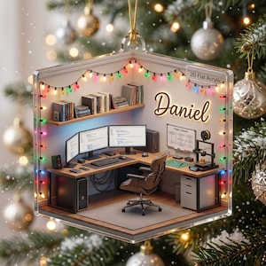 May include: A flat acrylic Christmas ornament featuring a miniature office scene with a desk, computer monitors, a chair, bookshelves, and a 3D printer. The wall displays the name Daniel and a project diagram, decorated with colorful string lights.