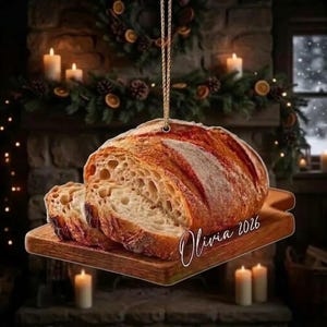 May include: A hanging ornament shaped like a wooden cutting board with a loaf of sliced sourdough bread on top. The bread has a golden-brown crust and an airy interior. The text Olivia 2026 is printed in white cursive script on the board.