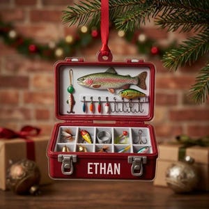 May include: A red tackle box Christmas ornament featuring the name ETHAN printed on the front. The open box displays a rainbow trout, various colorful fishing lures, hooks, and coiled line inside organized compartments, suspended by a red ribbon.