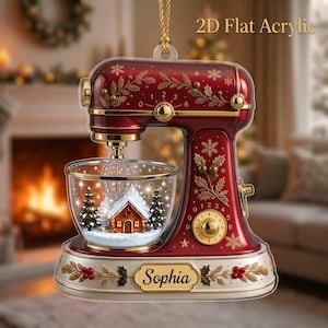 May include: A 2D flat acrylic ornament shaped like a red stand mixer with gold accents. The mixer features a clear bowl containing a miniature snowy house and pine trees. The base displays the name Sophia, and the text 2D Flat Acrylic is visible.