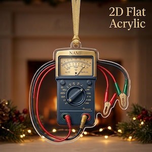 May include: A 2D flat acrylic hanging ornament shaped like a multimeter with a gold top section labeled NAME, a dark blue body, and red and black test leads with alligator clips. The gauge face displays Joy (V) and Spirits (A) with a star.