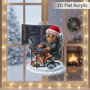 May include: A 2D flat acrylic ornament featuring a gingerbread person wearing a red Santa hat with the text YOUR NAME, a grey hoodie, and glasses. The character sits in a gaming chair at a desk with a laptop, monitor, and small gift boxes.