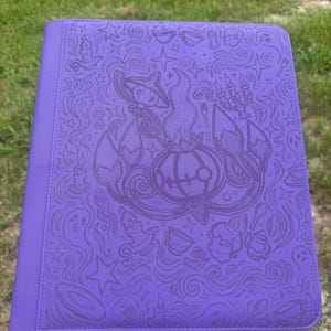 Peut inclure: Un classeur rectangulaire violet orné d'un motif en relief complexe représentant divers éléments fantaisistes, notamment un personnage rond central, des étoiles, des bougies, des tasses, des fantômes et des couverts.
